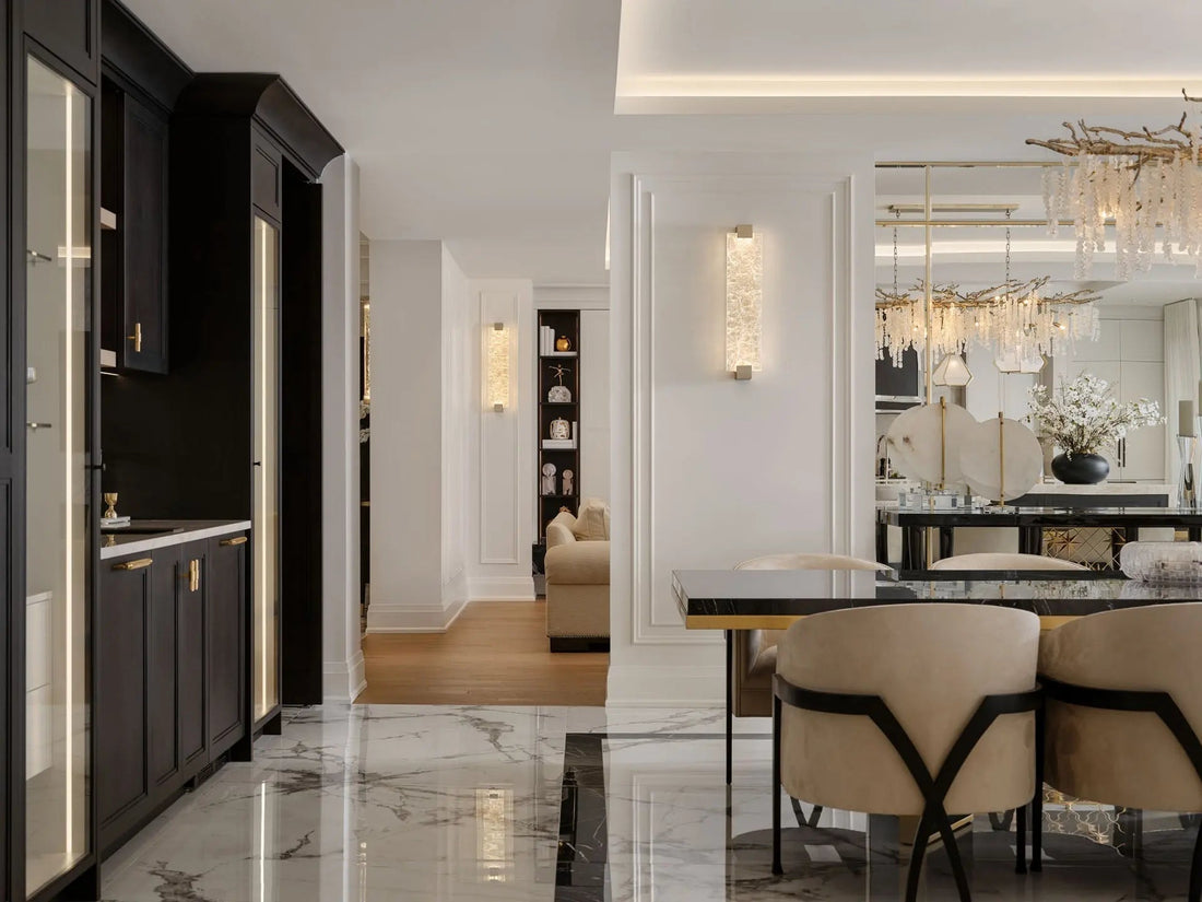 A lit crystal and nickel wall sconce glows on a white panelled column in an open-plan interior, with illuminated glass display cabinets in dark wood at left and the branch crystal chandelier over the dining table beyond.