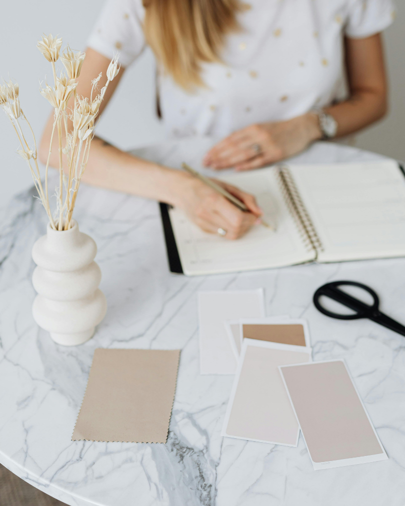 Person sketching or taking notes in a notebook at a marble table with neutral color swatches and styling materials.