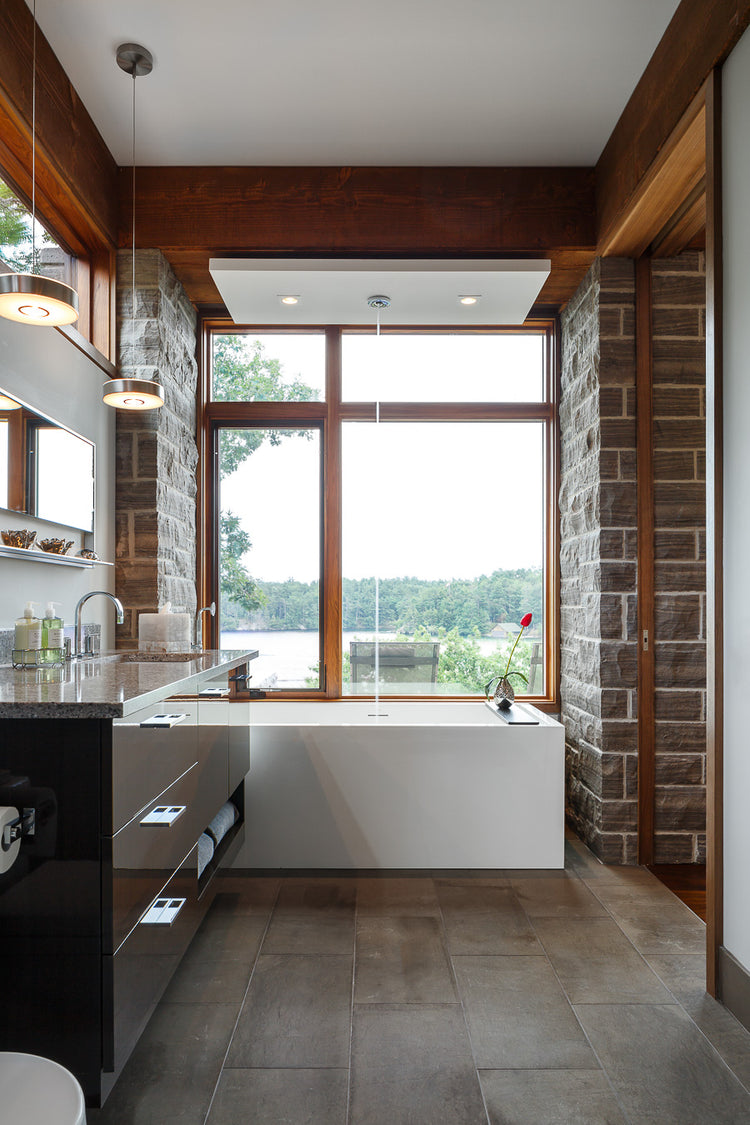 Modern bathroom with a soaking tub beneath a large window overlooking water, framed by wood beams and stone walls.