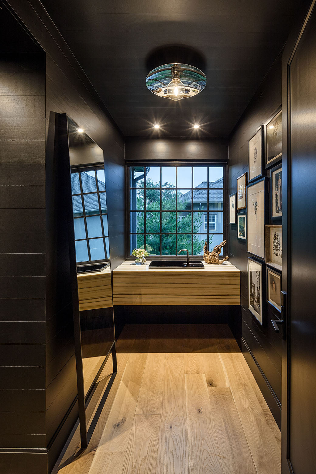 Moody modern bathroom with dark walls, wood vanity, framed art, and a window overlooking greenery.