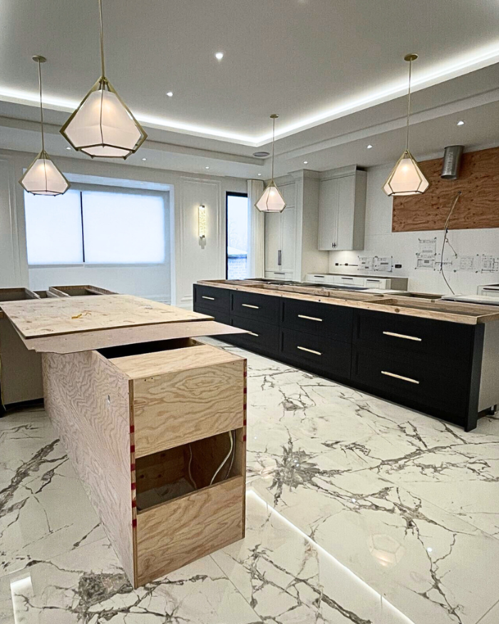 Modern kitchen under construction with large island cabinetry, marble-look flooring, pendant lighting, and unfinished wood panels and surfaces.
