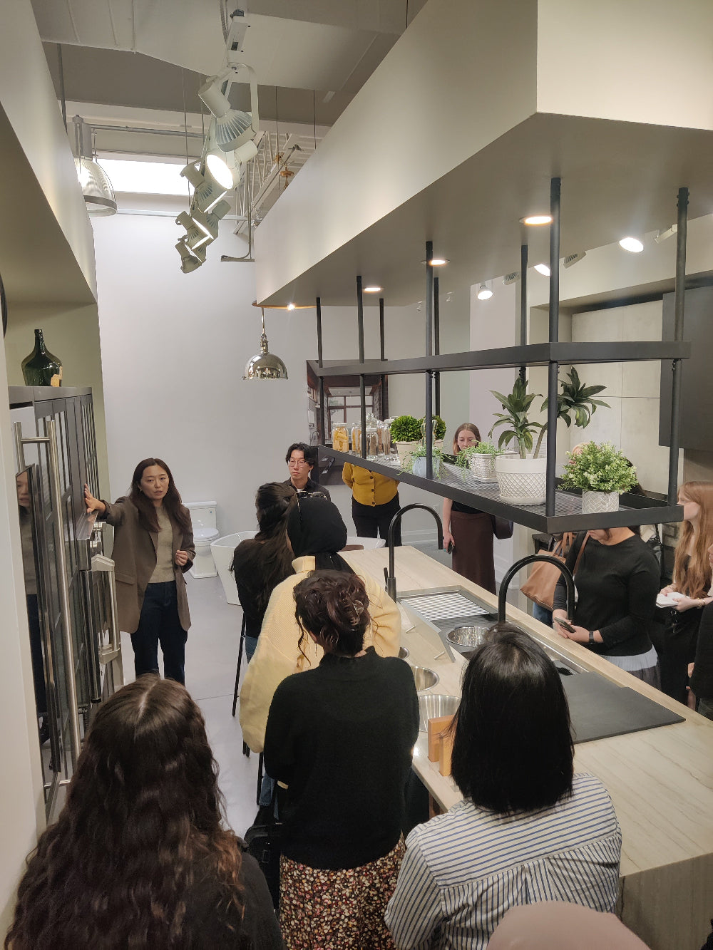 Group of people gathered in a modern kitchen showroom while a presenter explains features near cabinetry and appliances.