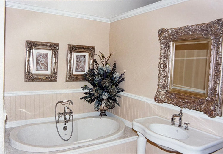 Traditional bathroom with a built-in tub, pedestal sink, ornate framed mirror, and decorative wall art.
