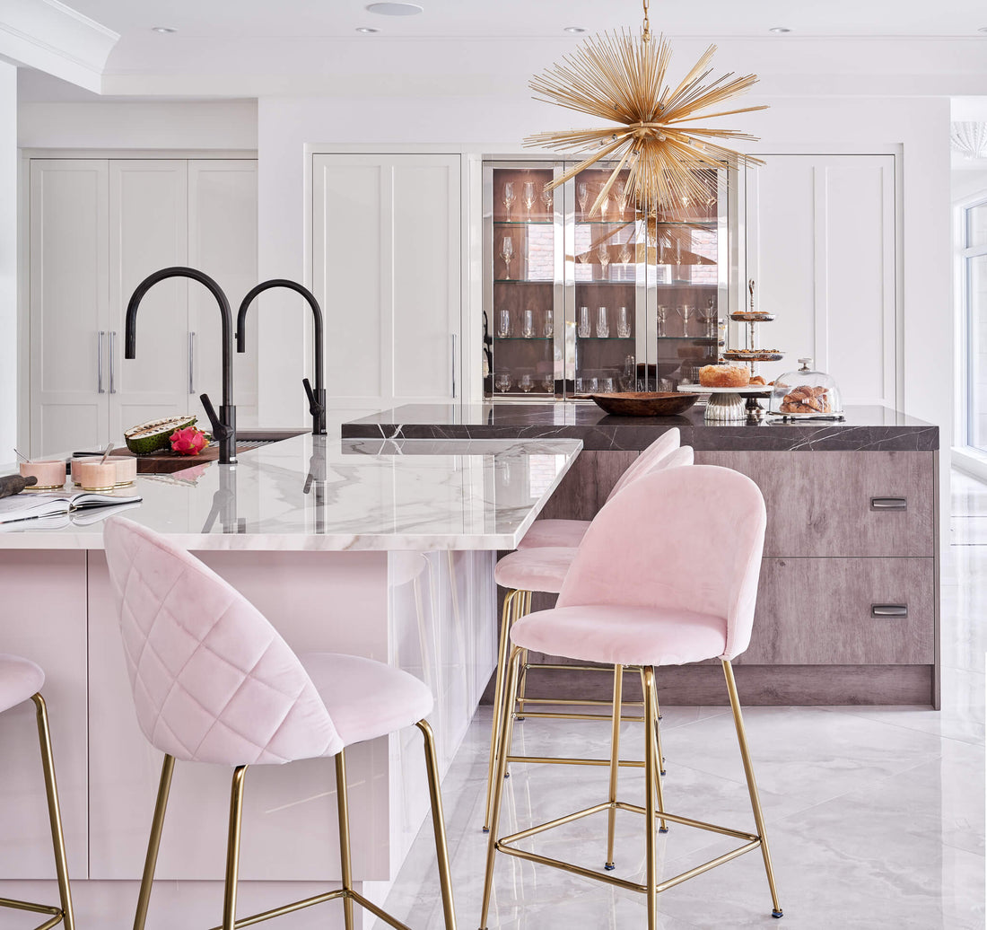 Elegant kitchen with marble island, pink upholstered bar stools, gold accents, and a statement chandelier.